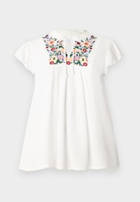 Blouse en coton blanc avec des manches courtes bouffantes, ornée de motifs floraux colorés brodés sur le plastron et d'un lien au niveau du col.
