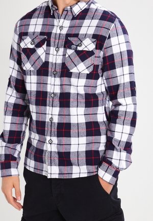 Muž v dlouho rukávové kostkované košili ve tmavě modré, bílé a červené barvě s dvěma náprsními kapsami a černých kalhotách s jednou rukou v kapse.