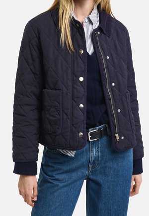 Femme portant une veste matelassée bleu marine avec des boutons-pression argentés et une fermeture éclair, superposée à un pull bleu marine et une chemise à rayures, associée à un jean bleu.