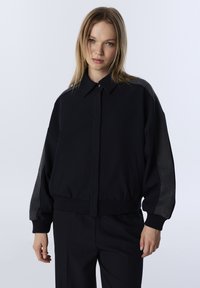 Zwarte bomberjack met een relaxte pasvorm, knoopsluiting bij de kraag en contrasterende grijze mouwen. Voorzien van zijzakken en geribbelde manchetten.