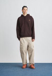 Felpa marrone con cappuccio, tasca a marsupio e logo ricamato, abbinata a pantaloni cargo beige con tasche laterali e calzature arancioni.