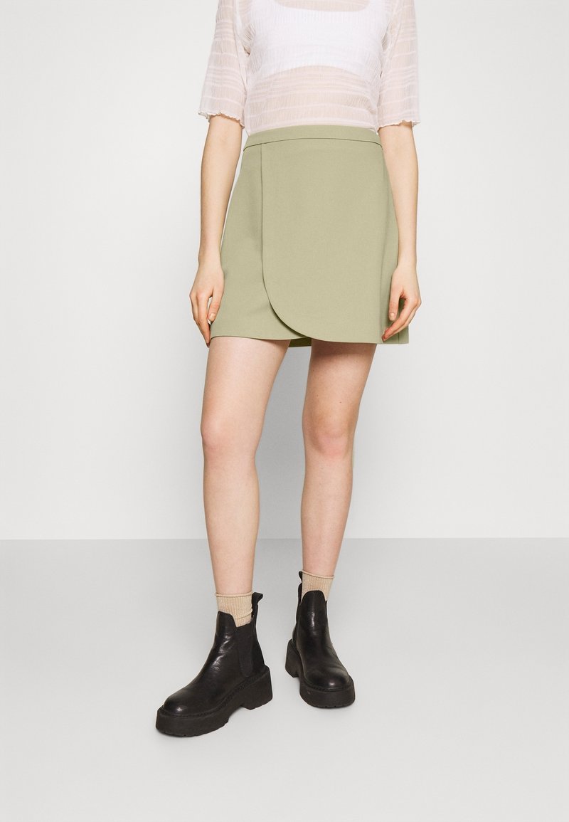 Vue sur toute la longueur des jambes d’une personne portant une mini-jupe portefeuille vert clair, un top blanc transparent, des chaussettes beiges et des bottines noires à plateforme.