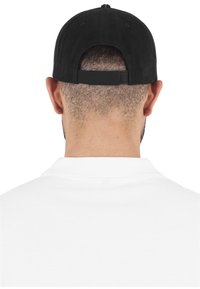 Svart caps med strukturert design, justerbar stropp og kontrasterende sømmer, båret med en hvit polo-skjorte. Vist fra baksiden.