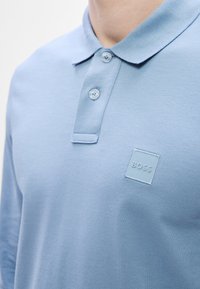 Šviesiai mėlynas polo marškinėlis su tekstūruota medžiaga, klasikiniu apykakliu ir dviem mygtukais, ant krūtinės turintis kvadratinį siuvinėtą logotipą.