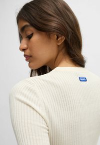 Pullover a coste color crema con maniche lunghe, scollatura rotonda e logo blu "HUGO" ricamato sulla schiena.