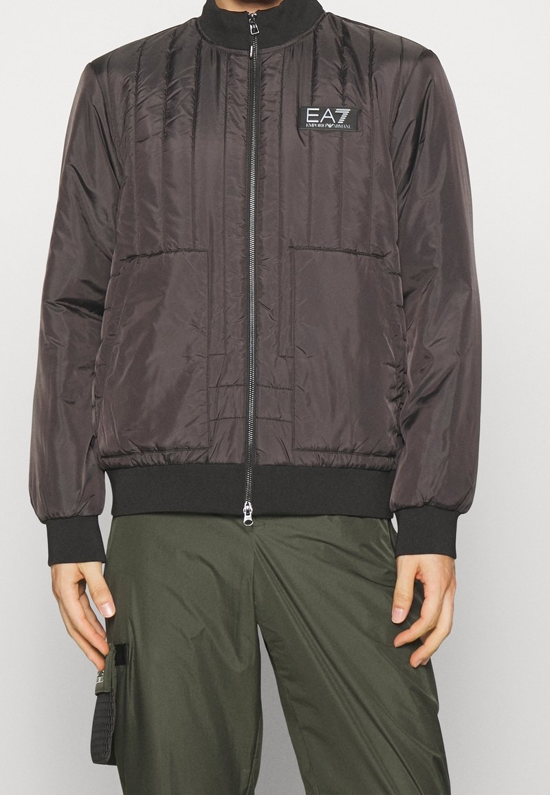Homme portant une veste matelassée noire zippée arborant le logo EA7 Emporio Armani et un pantalon cargo vert olive sur un fond uni.