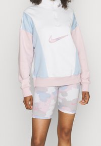 Pull-over Nike en blanc, bleu clair et rose, avec une fermeture à quart de zip et un logo. Assorti à un short de vélo avec un motif camouflage rose et bleu.