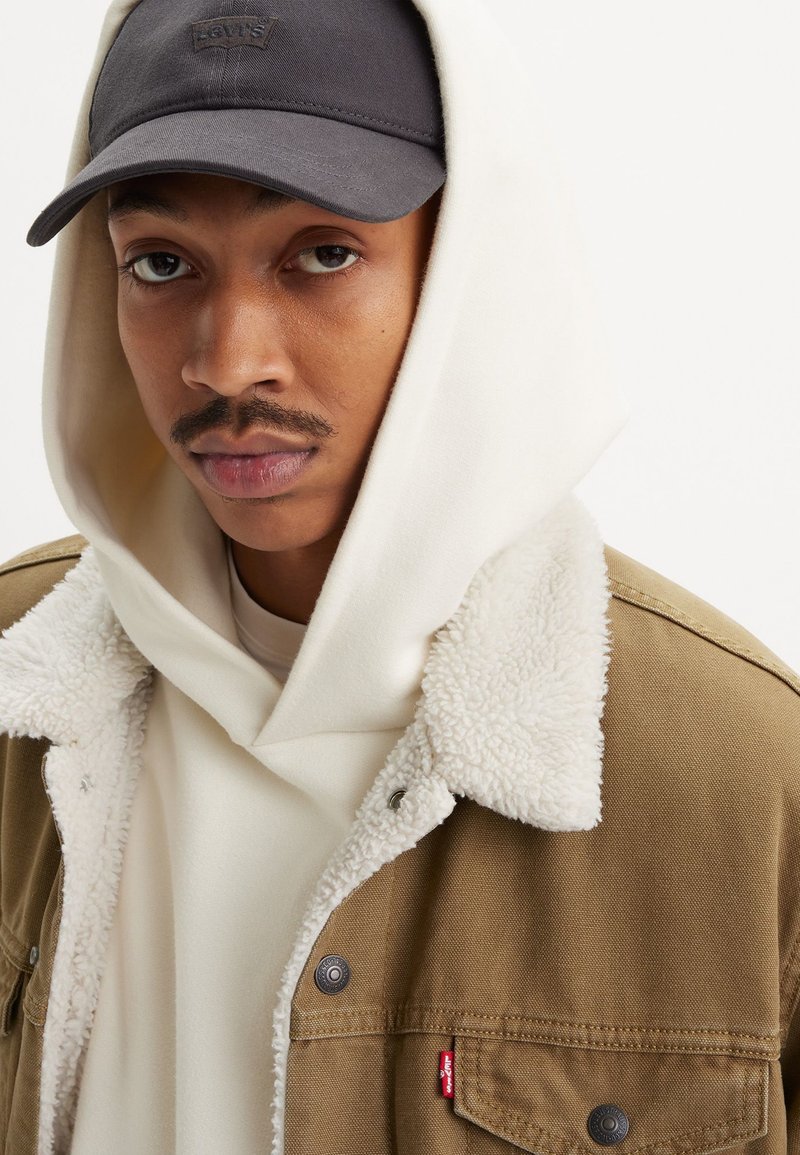 Veste marron avec doublure en polaire blanche, portée sur un sweat à capuche crème. Casquette noire avec logo sur le dessus. Gros plan, axé sur le visage et les détails du haut.