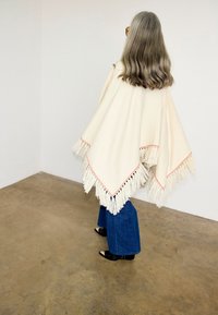 Γυναίκα με μακριά γκρι μαλλιά που φοράει εκρού πογκά με κρόσσια, μπλε παντελόνι φαρδιές παντελόνες και μαύρα παπούτσια με μυτερή μύτη, στέκεται σε τσιμεντένιο δάπεδο.