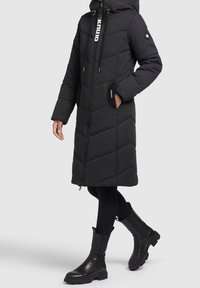 Abrigo acolchado negro con un patrón en forma de chevrón, capucha, cierre de cremallera y detalles de logo. Combinado con leggings negros y botas de tobillo. Textura suave.