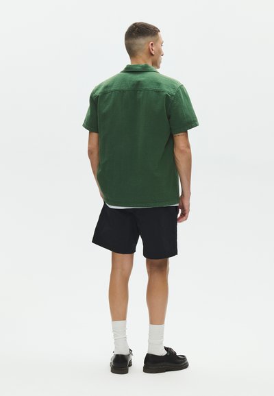 Homme de dos portant une chemise verte à manches courtes, un short noir, des chaussettes blanches et des chaussures noires sur un fond blanc uni.