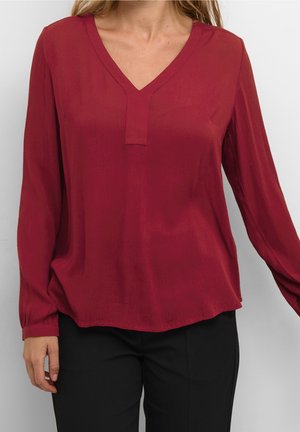 Kobieta w długim rękawie, ciemnoczerwonej bluzce z dekoltem w serek, w połączeniu z czarnymi spodniami na jednolitym tle.