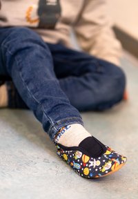 Stoff-Slipper mit dunklem Hintergrund und bunten Mustern von Planeten und Raumschiffen, weicher Sohle und elastischem Oberband, getragen mit Jeans.