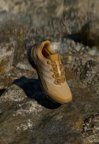 Brązowe buty outdoorowe z teksturowanej tkaniny i gumową podeszwą, z zielonymi akcentami, sznurówkami i trzema białymi paskami z boku.