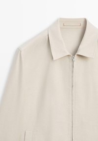 Beige Reißverschlussjacke mit Kragen und langen Ärmeln, mit einer Taschenverzierung auf der linken Seite.