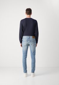 Camisa de manga larga ajustada de color azul marino para hombre, combinada con jeans de denim azul claro. Los jeans cuentan con un parche de cuero y cinco bolsillos. Zapatillas blancas completan el look.