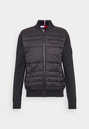 Schwarze Jacke mit Reißverschluss, gesteppte Vorderseite, glatte Ärmel, zwei Druckknopftaschen und kleines rot-weiß-blaues Logo auf der Brust.