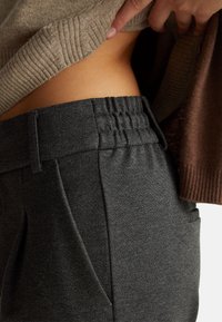 Šedé elastické kalhoty s elastickým pasem, bočními kapsami a hladkým povrchem, v kombinaci s lehkým pleteným topem a hnědými akcenty.