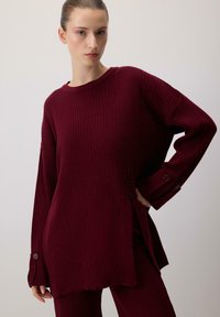 Bordowy sweter o prążkowanej strukturze, z obniżonymi ramionami, rozcięciem po bokach i guzikami na mankietach. Miękka tekstura, luźny krój i długie rękawy.