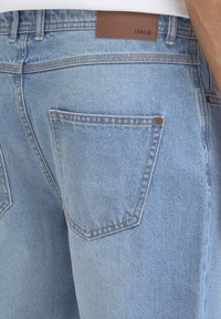 Hele sinine teksapüksid, millel on viie taskuga disain, pruun nahast silt vöökohal ja kontrastne õmblus õmbluste ääres.