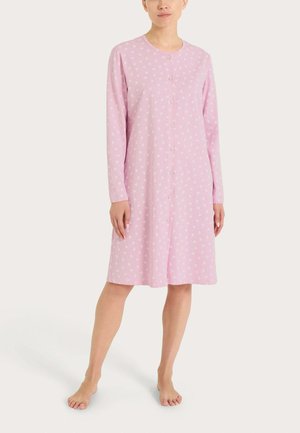 Donna che indossa un camicione da notte rosa, lungo fino al ginocchio, con maniche lunghe, piccoli motivi floreali bianchi e bottoni frontali, in piedi a piedi nudi.