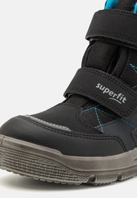 Zwarte kinderschoen met een gestructureerde bovenkant, klittenbandsluitingen en blauwe stiksels. Dikke rubberen zool met ribbels voor grip. Merkennaam zichtbaar.