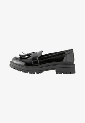 Schwarzer Lackleder-Slipper mit klobiger Sohle und Quasten-Detail oben, Seitenansicht vor weißem Hintergrund.