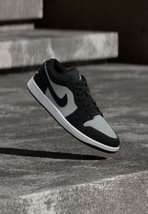 Čierno-šedá teniska Nike vznášajúca sa nad textúrovaným betónovým povrchom s kamennými schodmi v pozadí.