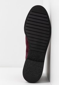 Semelle d'une chaussure violet foncé avec une semelle extérieure en caoutchouc noir texturé présentant des rainures horizontales et une surface de talon rugueuse, inclinée contre un coin blanc.