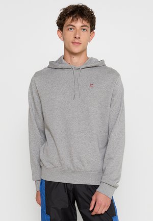 Young man with short curly brown hair wearing a light gray hooded sweatshirt and black pants with blue panels standing against a plain background.