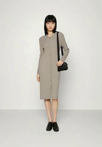 Mulher vestindo um vestido midi bege com botões, bolsa preta a tiracolo e sapatos pretos, de pé contra um fundo claro e liso.