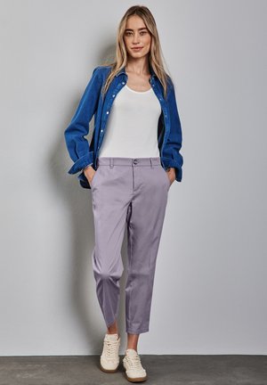 Helllila Hosen in schmaler Passform, kombiniert mit einem weißen Tanktop und einem blauen Jeanshemd, getragen mit cremefarbenen Sneakers. Glatte Stoffoberfläche.