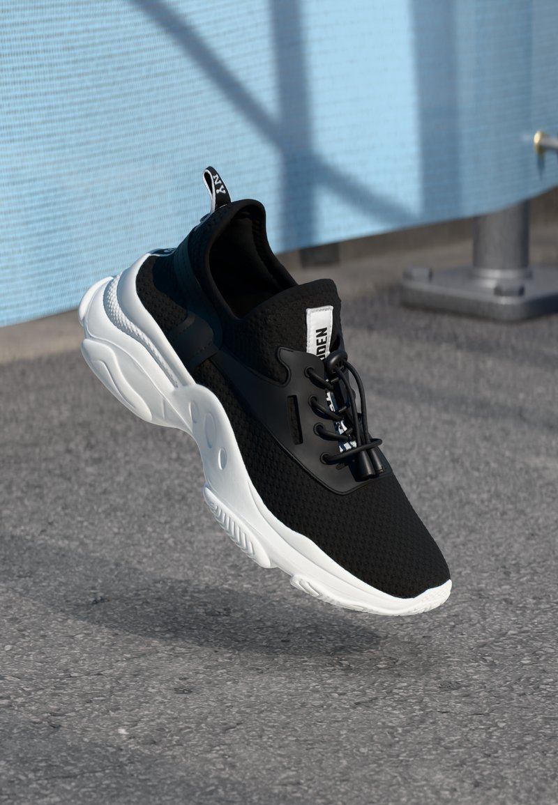 Black and white athletic shoe with mesh upper and thick sole, suspended above textured concrete surface with a blue textured wall background.