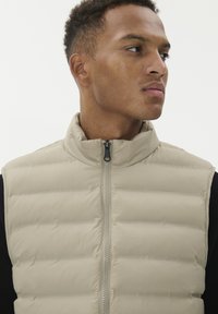 Mees, kes kannab beeži nööriga magusat ilma varrukat, vaadates paremale valge tausta taustal.