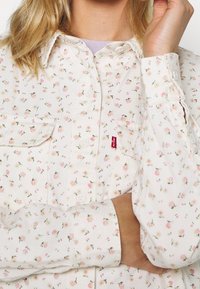 Chemise à boutons à imprimé floral sur fond blanc, avec de petites fleurs roses, deux poches poitrine et une étiquette du logo Levi's sur le côté.