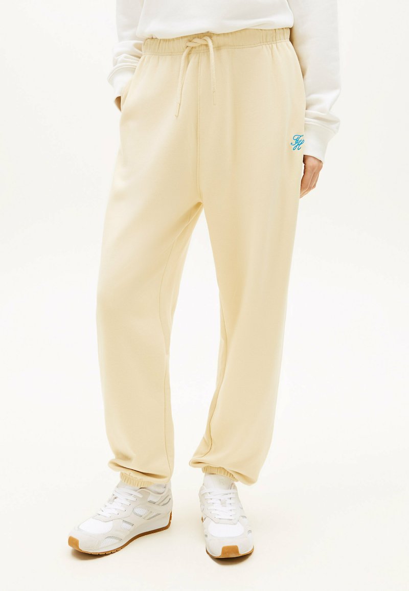 Pantalon de survêtement beige avec taille et chevilles élastiques, logo bleu sur la cuisse, porté avec des baskets blanches et un haut à manches longues blanc.
