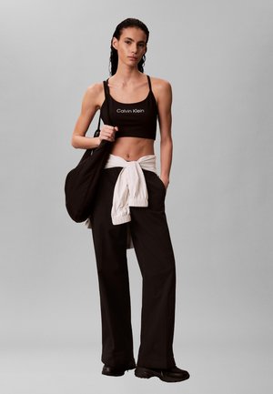 Mladá žena v černém crop topu Calvin Klein, černých kalhotách s širokými nohavicemi, bílé bundě uvázané kolem pasu, černých teniskách a s černou taškou.