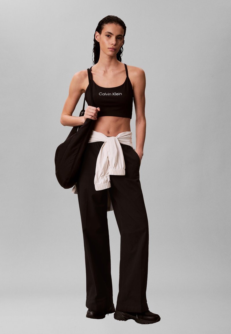 Νεαρή γυναίκα που φορά μαύρο crop top Calvin Klein, μαύρο παντελόνι φαρδύ, λευκό μπουφάν δεμένο γύρω από τη μέση, μαύρα αθλητικά παπούτσια και κρατά μαύρη τσάντα.