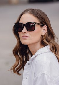 Černé oversized sluneční brýle s tmavými skly, s geometrickým rámem. Modelka má na sobě bílou bundu s límečkem a malé zlaté náušnice.