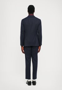 Uomo in piedi di spalle, vestito con un abito navy a righe, calze bianche e scarpe nere, su uno sfondo chiaro e semplice.