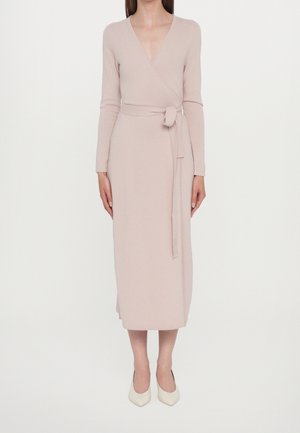 Femme portant une robe portefeuille beige à manches longues avec une ceinture en tissu nouée à la taille et des chaussures blanches à bouts pointus.