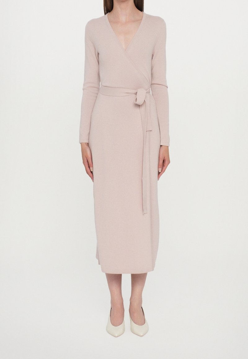 Femme portant une robe portefeuille beige à manches longues avec une ceinture en tissu nouée à la taille et des chaussures blanches à bouts pointus.