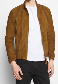 Brun mockajacka med dragkedja fram, långa ärmar och synliga sömdetaljer. Bärs över en vit t-shirt och matchas med mörka jeans.