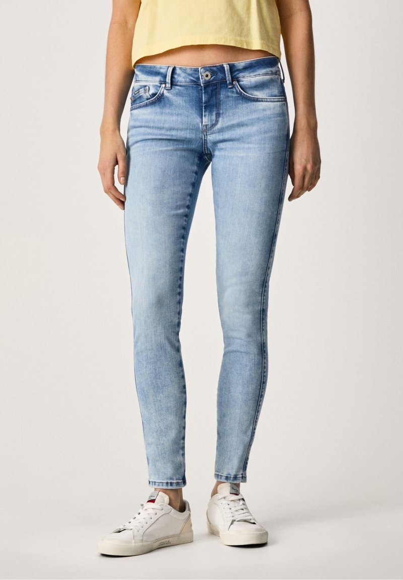 Vaqueros skinny de un azul claro deslavado, diseño de cinco bolsillos, con cierre de cremallera y botón, combinados con zapatillas blancas. Textura de denim suave.