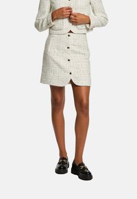 Valkoinen tweed mini-hame napituksella edessä, jossa on hienovarainen tekstuurikuvio ja halkio, yhdistetty mustiin paksupohjaisiin loafer-kenkiin.