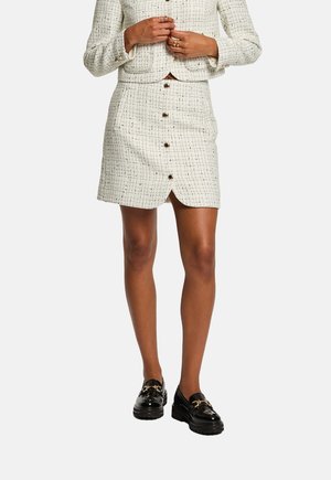 Valkoinen tweed mini-hame napituksella edessä, jossa on hienovarainen tekstuurikuvio ja halkio, yhdistetty mustiin paksupohjaisiin loafer-kenkiin.