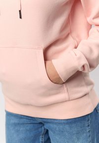 Svetlo roza kapa s kenguru žepom, rebrastimi manšetami in spodnjim robom. Mehka tkanina z gladko teksturo, prikazana z modrimi jeansi iz jeansa.