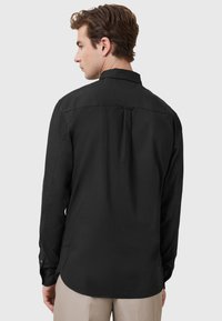 Mann mit braunen Haaren, der ein schwarzes Langarmhemd mit Knöpfen und beigefarbene Hosen trägt, von hinten vor einem schlichten Hintergrund gezeigt.