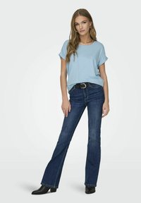 Lys blå kortermet topp med rullekanter, kombinert med mørke utsvingte jeans. Modellen har på seg et svart belte og ankelstøvler. Enkelt design.