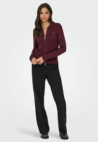 Sieviete stāv, uzvilkusi bordo krāsas adītu uzvalku ar rāvējslēdzēju un mazu sirds emblēmu, melnas platās bikses un tumšas kurpes gaišā fona priekšā.